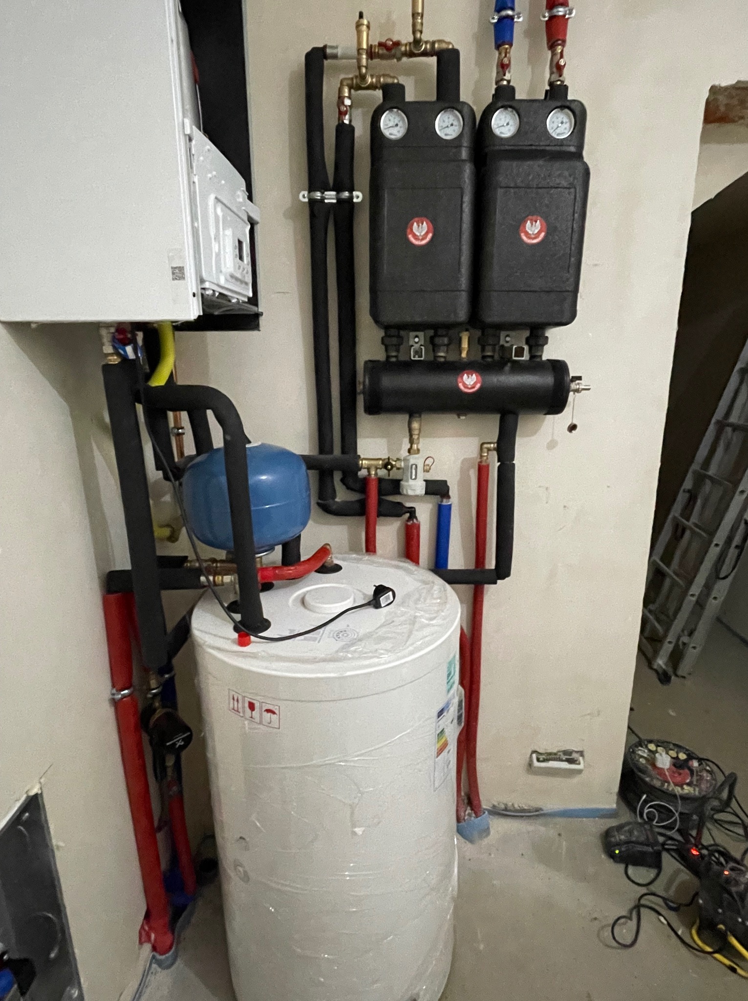 Nowoczesny system grzewczy w trakcie instalacji: biały zasobnik ciepłej wody owinięty folią, wiszący kocioł, czarne rury izolacyjne, czerwone i niebieskie rury doprowadzające wodę, manometry...