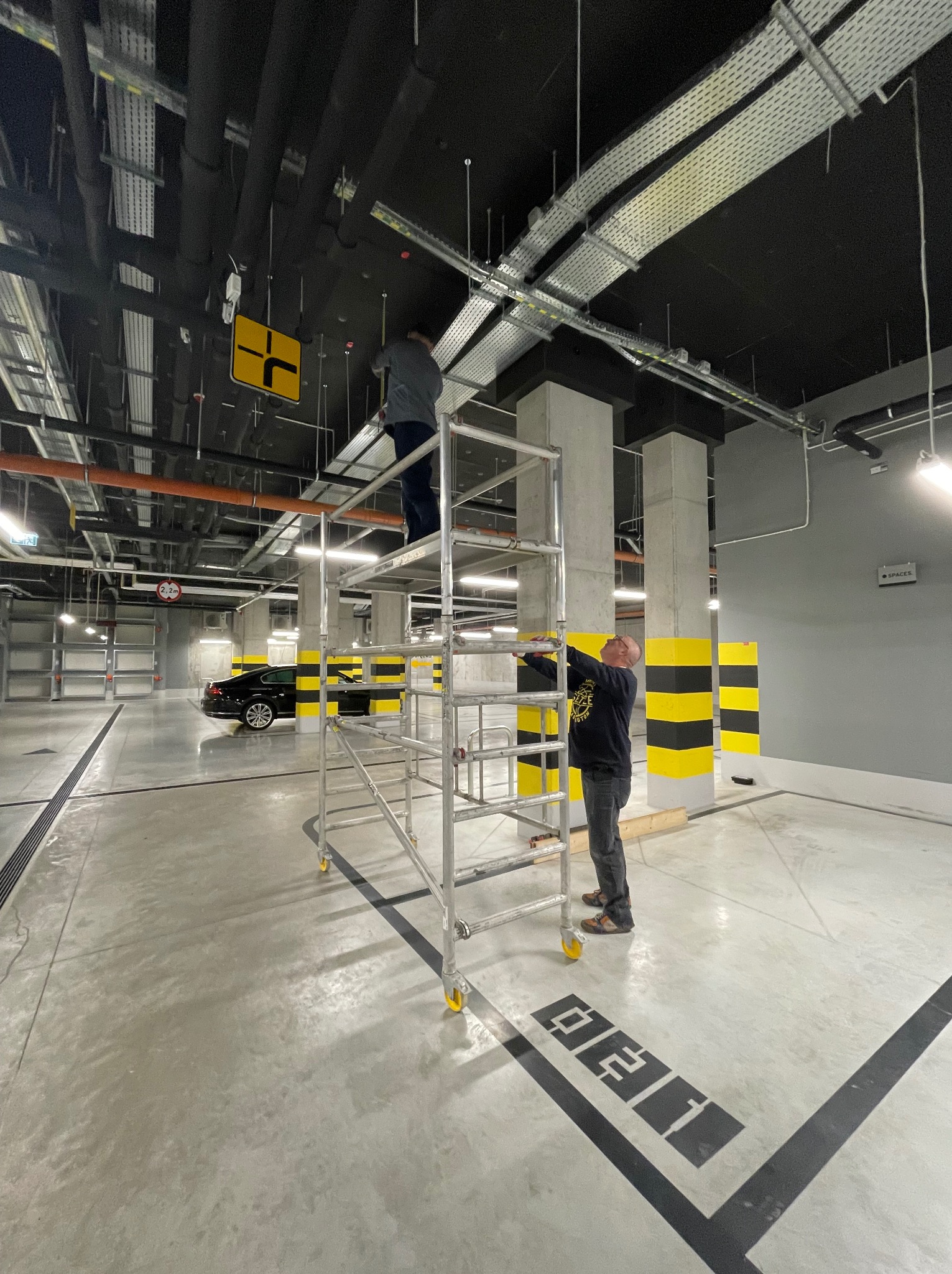 Dwóch mężczyzn na rusztowaniu w garażu podziemnym montuje instalacje pod sufitem, widoczne koryta kablowe i rury wentylacyjne.