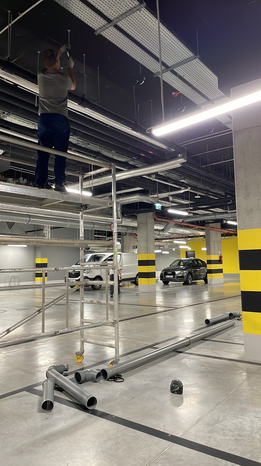 Instalator na rusztowaniu montuje rury pod sufitem parkingu podziemnego, widoczne elementy instalacji i narzędzia pomiarowe.