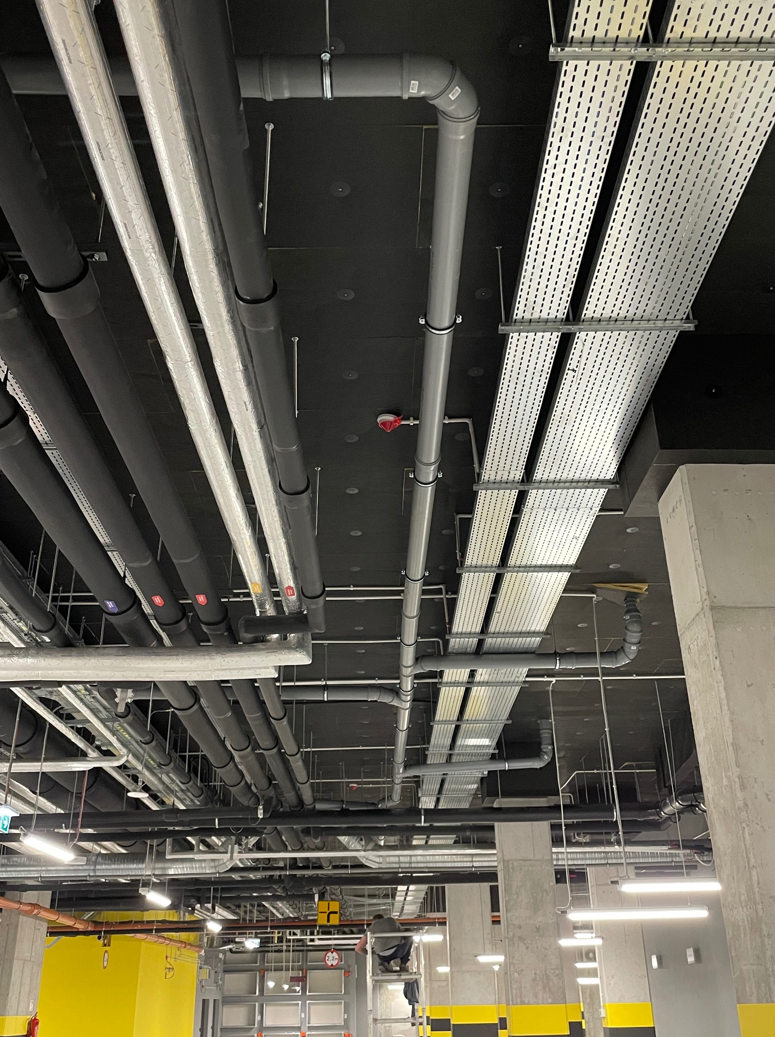 Widok instalacji pod sufitem w garażu podziemnym: rury hydrauliczne w izolacji, rury kanalizacyjne, koryta kablowe, oświetlenie, elementy wentylacji i system przeciwpożarowy.