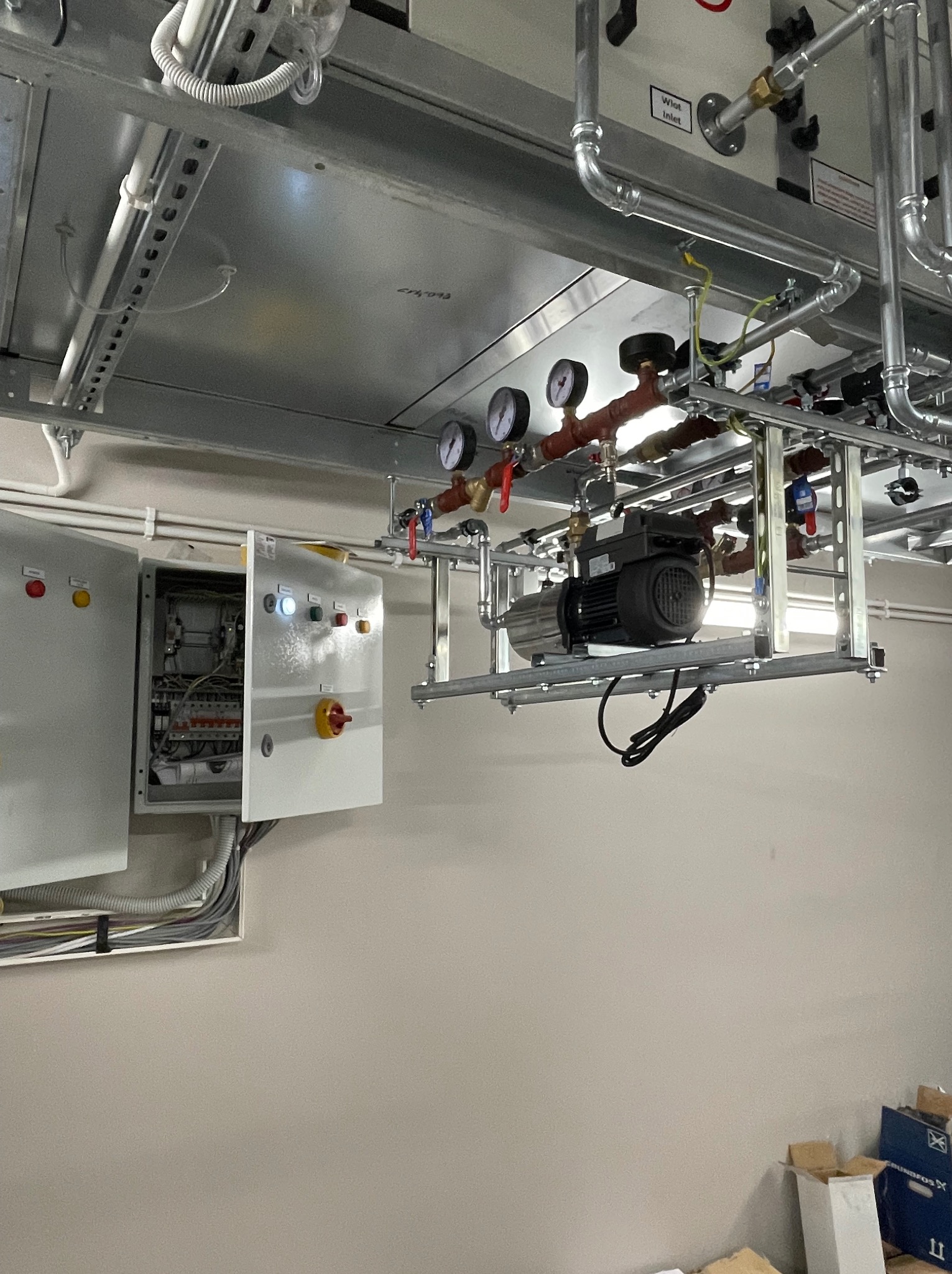Widok na zawieszoną instalację hydrauliczną z pompą obiegową, manometrami i rurami, oraz otwartą szafę sterowniczą na ścianie.