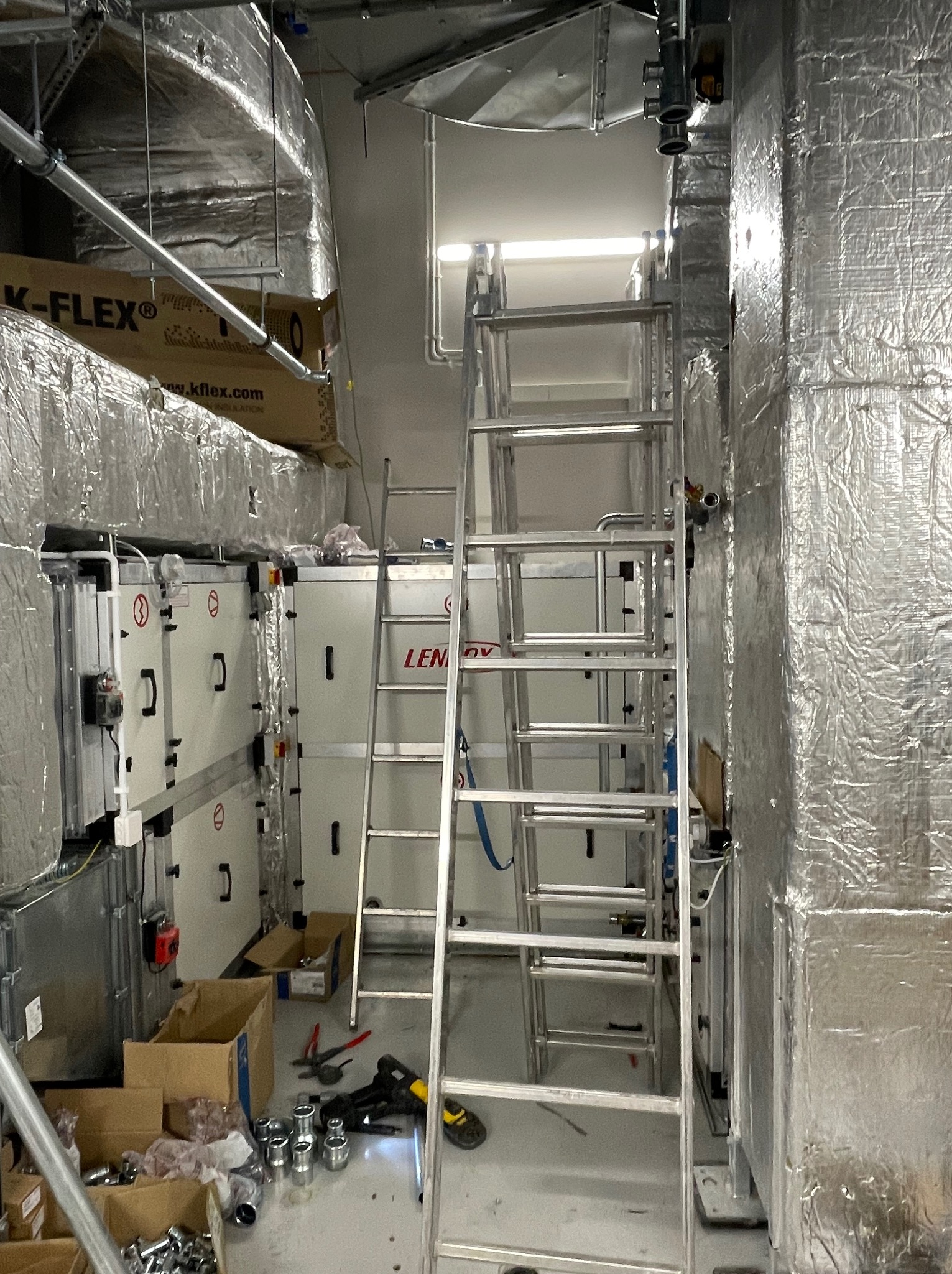 Wnętrze techniczne z instalacjami wentylacyjnymi i klimatyzacyjnymi w trakcie montażu, widoczne izolowane kanały, jednostki centralne oraz drabina aluminiowa.