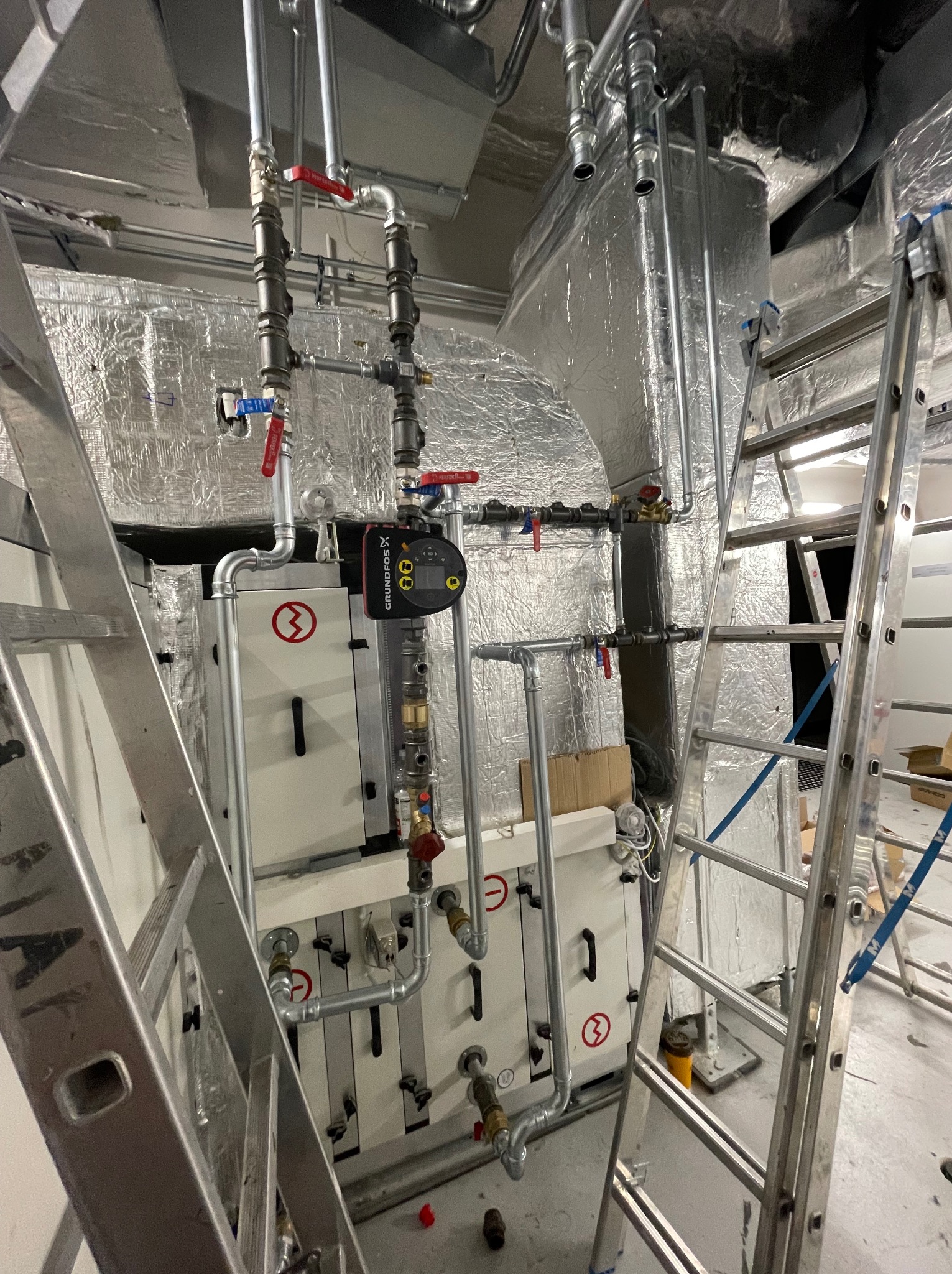 Instalacja grzewcza w kotłowni z widocznymi rurami, zaworami, pompą obiegową Grundfos i drabinkami aluminiowymi, izolacja termiczna na ścianie i suficie.