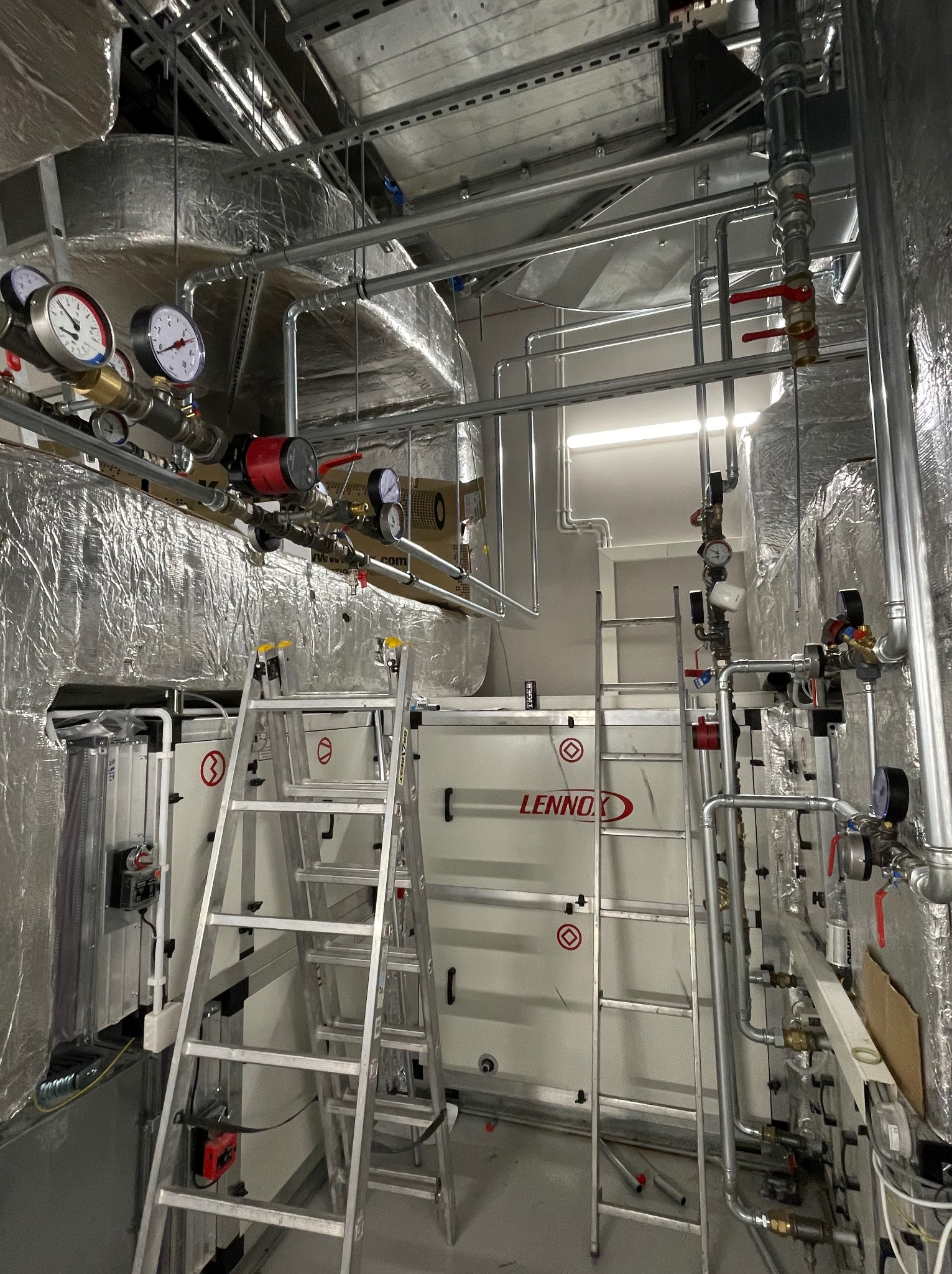 Rozbudowana instalacja grzewcza w pomieszczeniu technicznym, z widocznymi rurami, zaworami, manometrami, izolacją termiczną, drabina i centralą wentylacyjną Lennox.