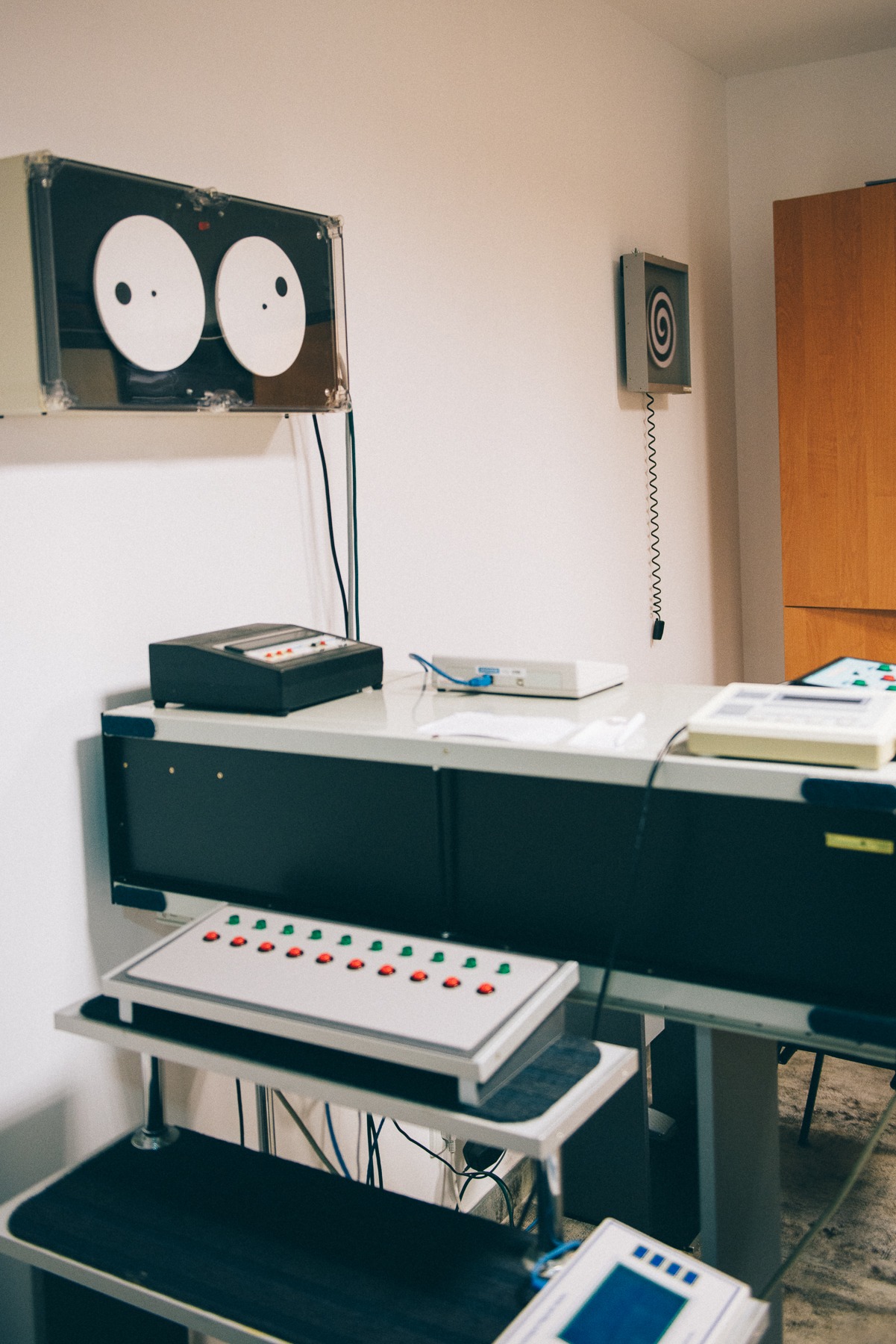 Aparatura diagnostyczna w gabinecie z dwoma okrągłymi ekranami z czarnymi punktami, panelem kontrolnym z czerwono-zielonymi przyciskami i spiralą na ścianie.