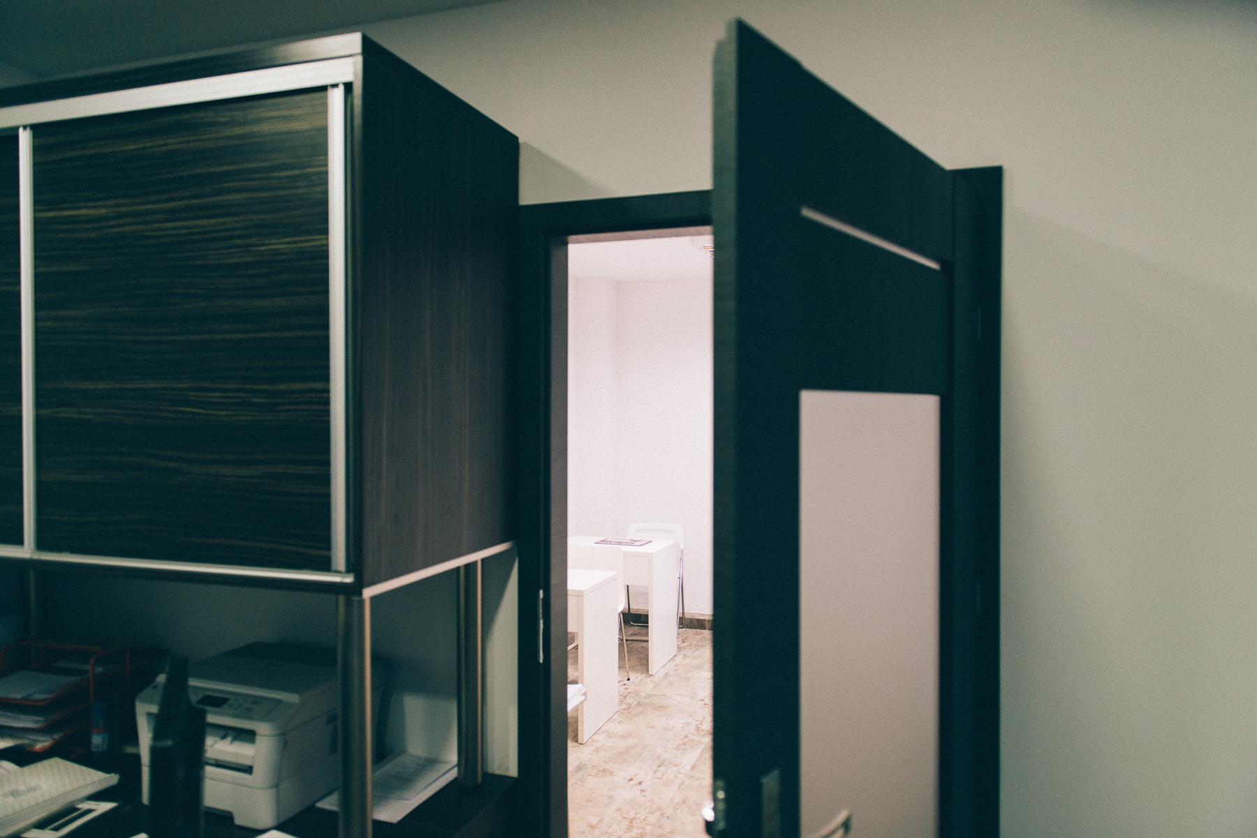 Wnętrze gabinetu z biurkiem i krzesłami widoczne przez otwarte drzwi, obok szafa z przesuwnymi drzwiami i drukarka na biurku.