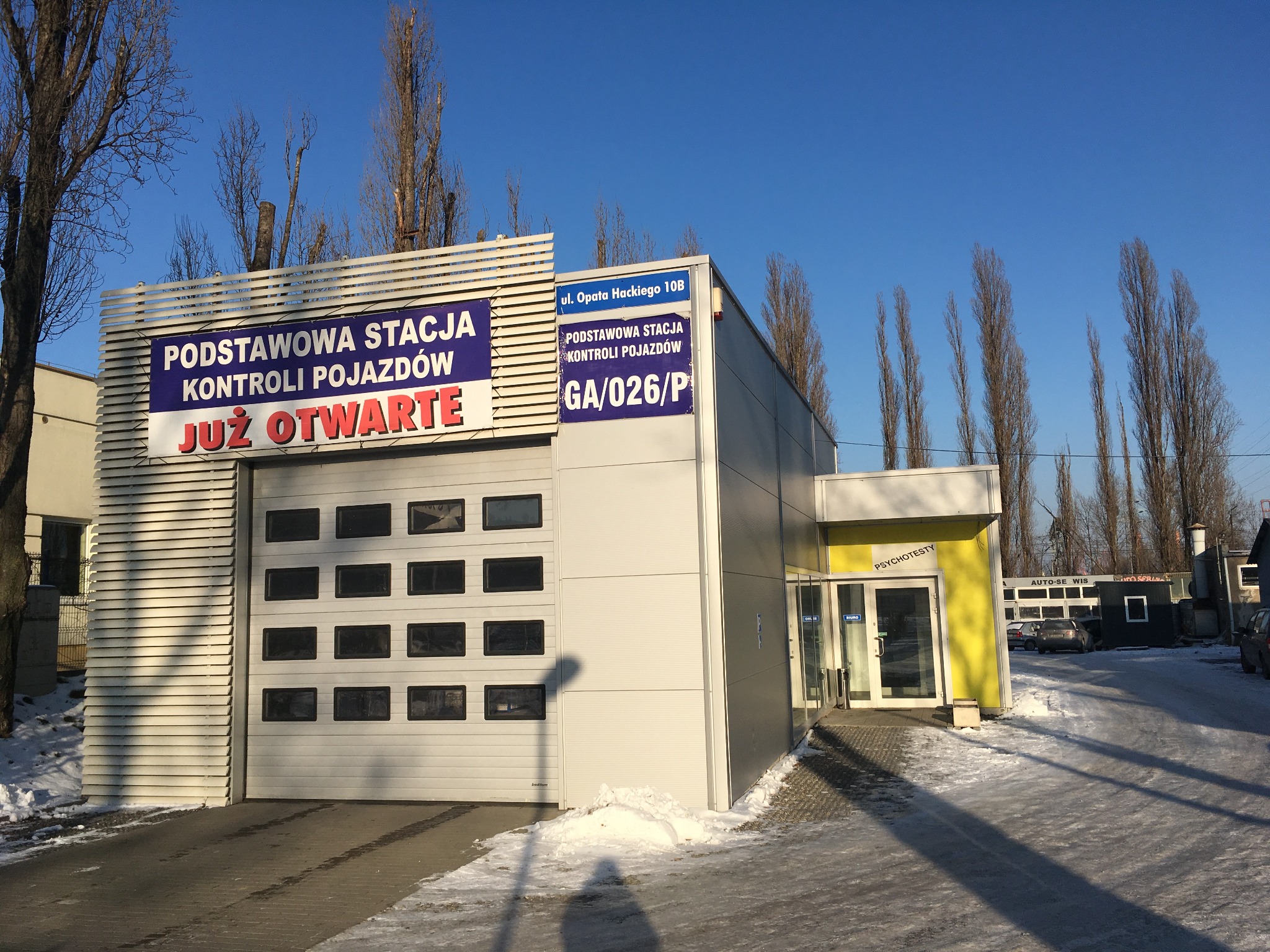 Budynek stacji kontroli pojazdów z napisem 'Podstawowa Stacja Kontroli Pojazdów JUŻ OTWARTE' oraz informacją o psychotestach na żółtej ścianie przy wejściu.