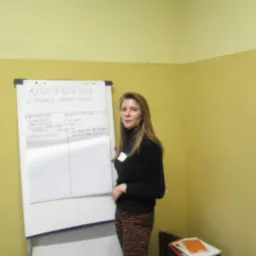 Kobieta w czarnym golfie i wzorzystych spodniach stoi obok flipcharta z napisem 'Myśli i działania w sytuacji utraty pracy', prezentując treści szkoleniowe w żółtym pomieszczeniu.