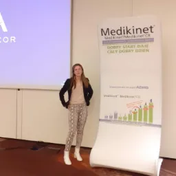 Kobieta w spodniach w wężowy wz&oacute;r stoi obok roll-upu reklamowego z informacjami o leku Medikinet i terapii ADHD.