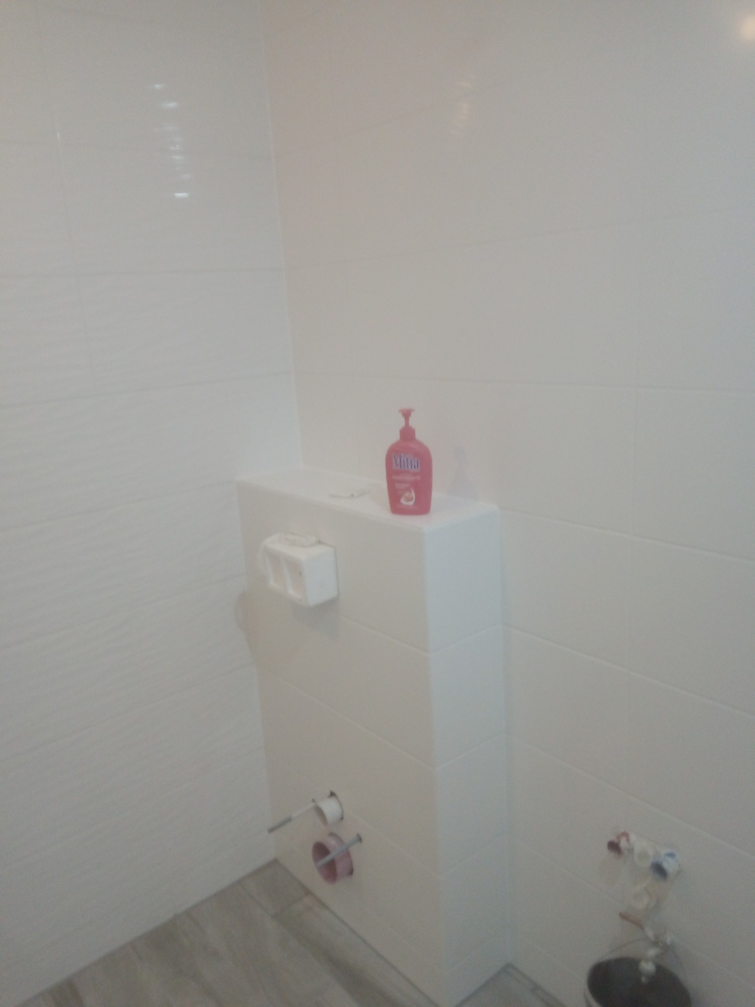 Biała łazienka z ukrytą spłuczką, wykończona płytkami imitującymi falę, z dozownikiem mydła w kolorze różowym na górze. Widoczne przyłącza wody i pojemnik na papier toaletowy.