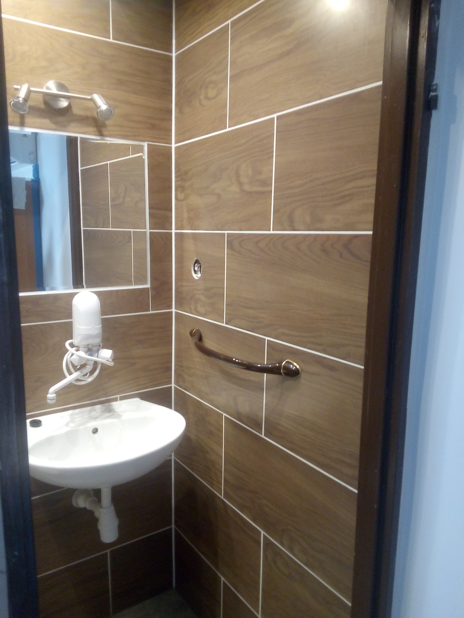 Umywalka zamontowana w rogu łazienki, obok poręcz dla niepełnosprawnych, ściany wyłożone brązowymi płytkami imitującymi drewno z jasnymi fugami, nad umywalką dozownik mydła.