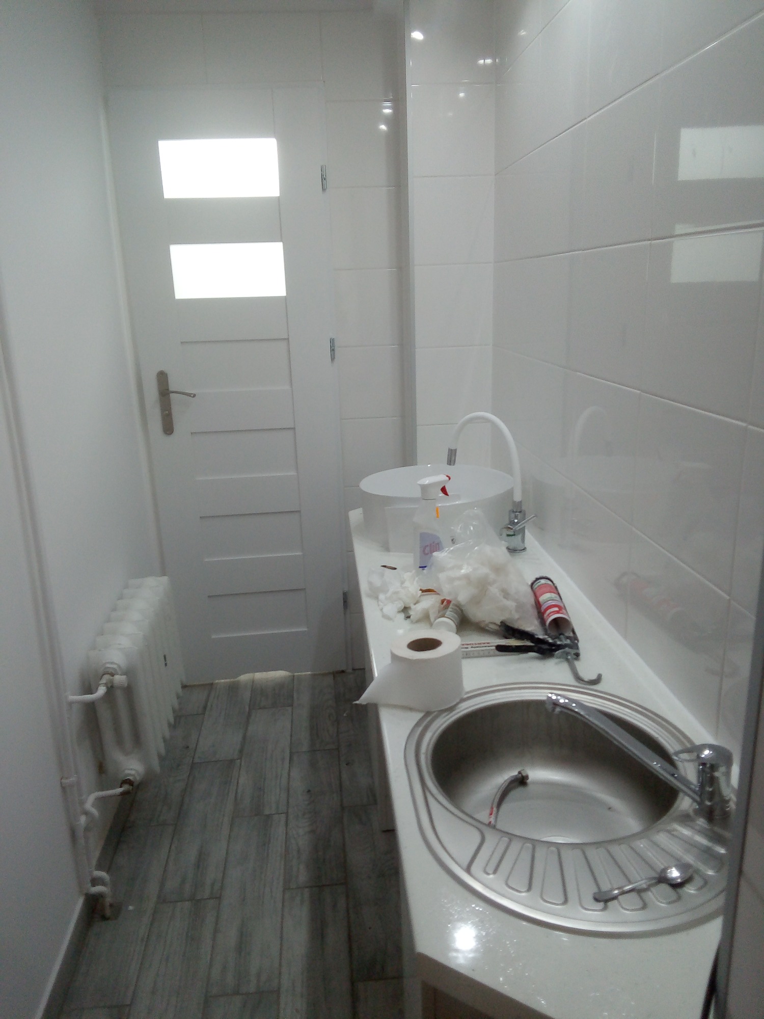 Biała łazienka w trakcie montażu umywalki: widoczna stalowa umywalka z baterią, biała szafka, białe płytki ścienne, narzędzia, silikon, papier toaletowy i grzejnik żeberkowy.