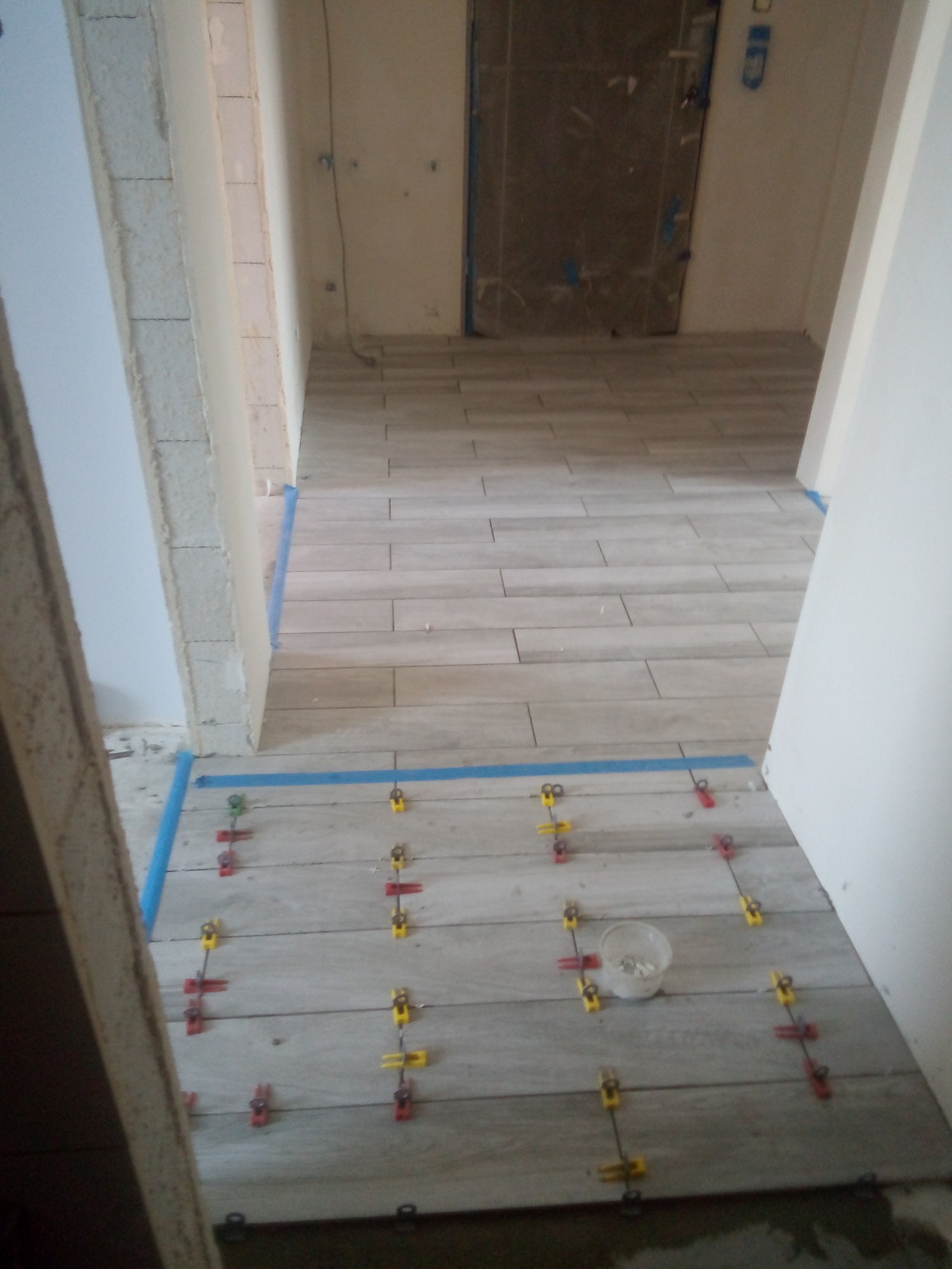 Układanie podłogowych płytek imitujących drewno, z użyciem poziomujących klipsów i niebieskiej taśmy malarskiej, widok na korytarz.