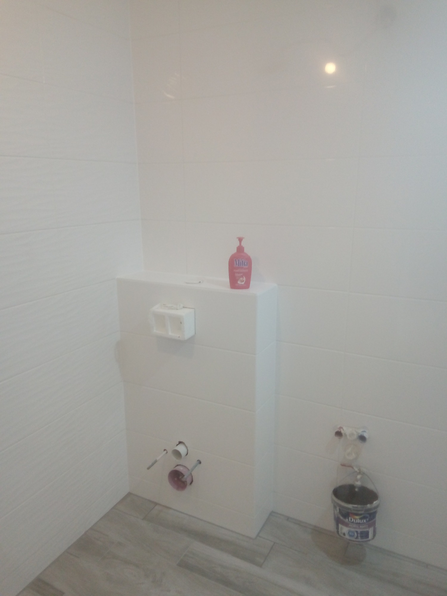 Biała łazienka z widoczną instalacją podtynkową, płytkami imitującymi drewno na podłodze, różowym mydłem w płynie na zabudowie oraz wiadrem z farbą Dulux, sugerującym prace wykończeniowe.
