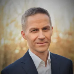 Portret mężczyzny w garniturze na rozmytym tle. Delikatny uśmiech, krótko ostrzyżone włosy. Profesjonalny wizerunek.