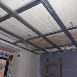 Montaż metalowej konstrukcji pod sufitem podwieszanym z widocznymi elementami mocującymi, instalacją elektryczną i fragmentem drabiny oparta o fioletową płytę gipsową.