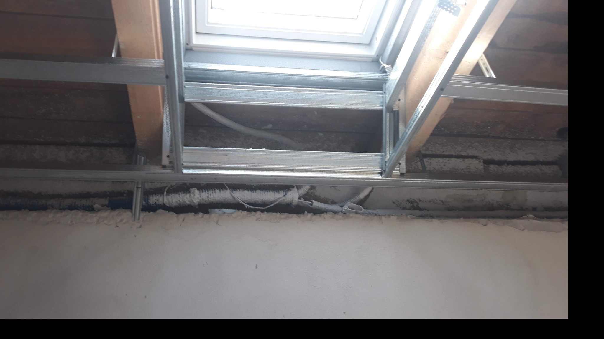 Metalowa konstrukcja sufitu podwieszanego z widocznym oknem dachowym i fragmentami instalacji elektrycznej w przestrzeni między sufitem a konstrukcją.