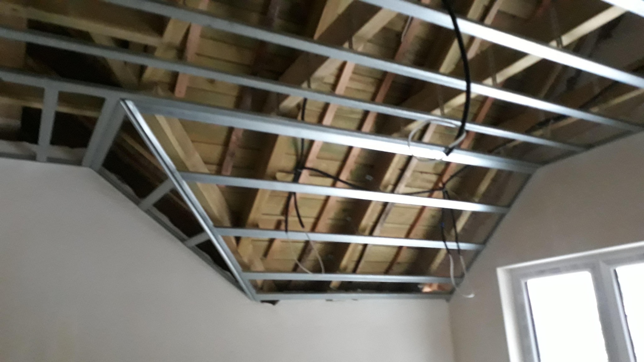 Metalowa konstrukcja sufitu podwieszanego z widocznymi przewodami elektrycznymi, zamontowana w pomieszczeniu ze skośnym sufitem i oknem.