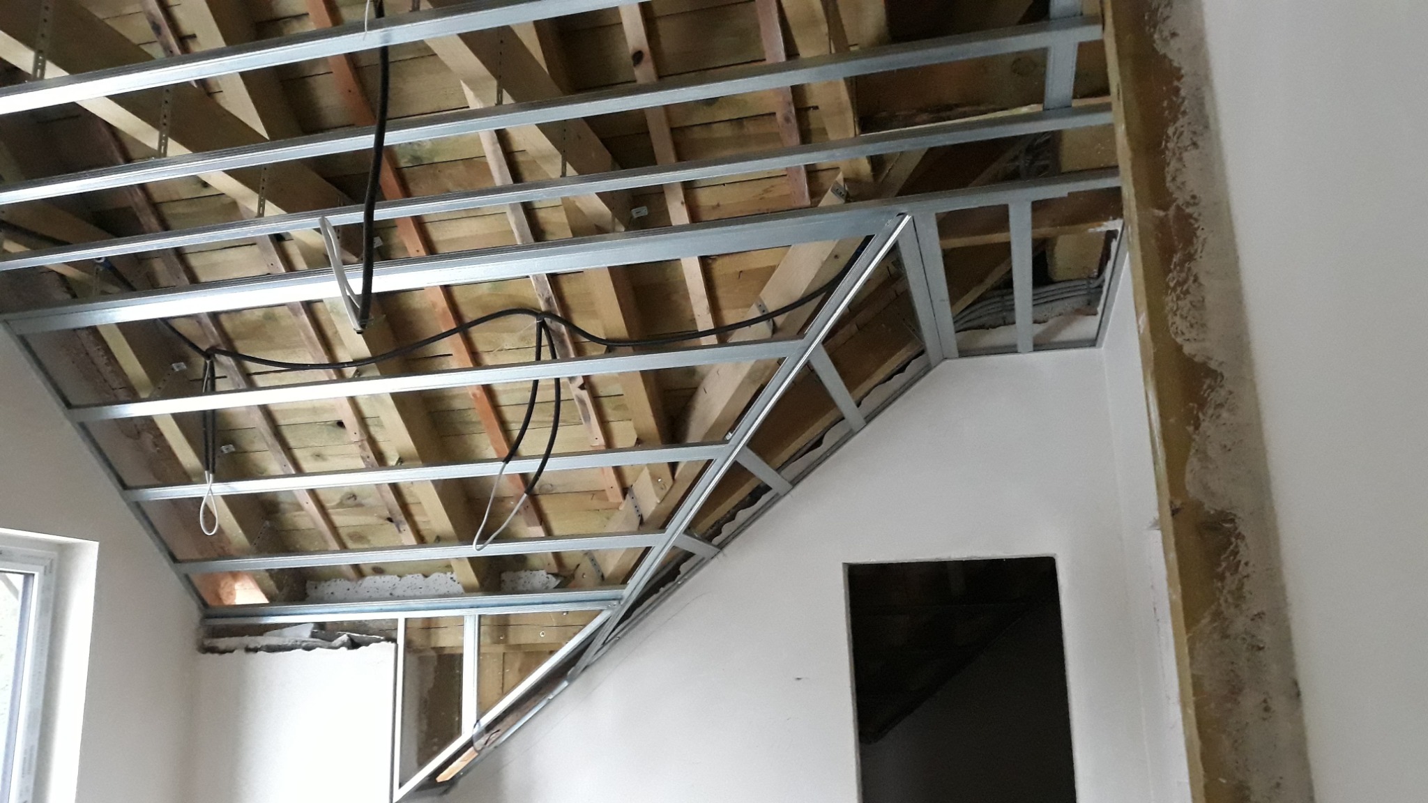 Montaż metalowej konstrukcji sufitu podwieszanego na poddaszu, widoczne drewniane elementy więźby dachowej i przewody elektryczne.