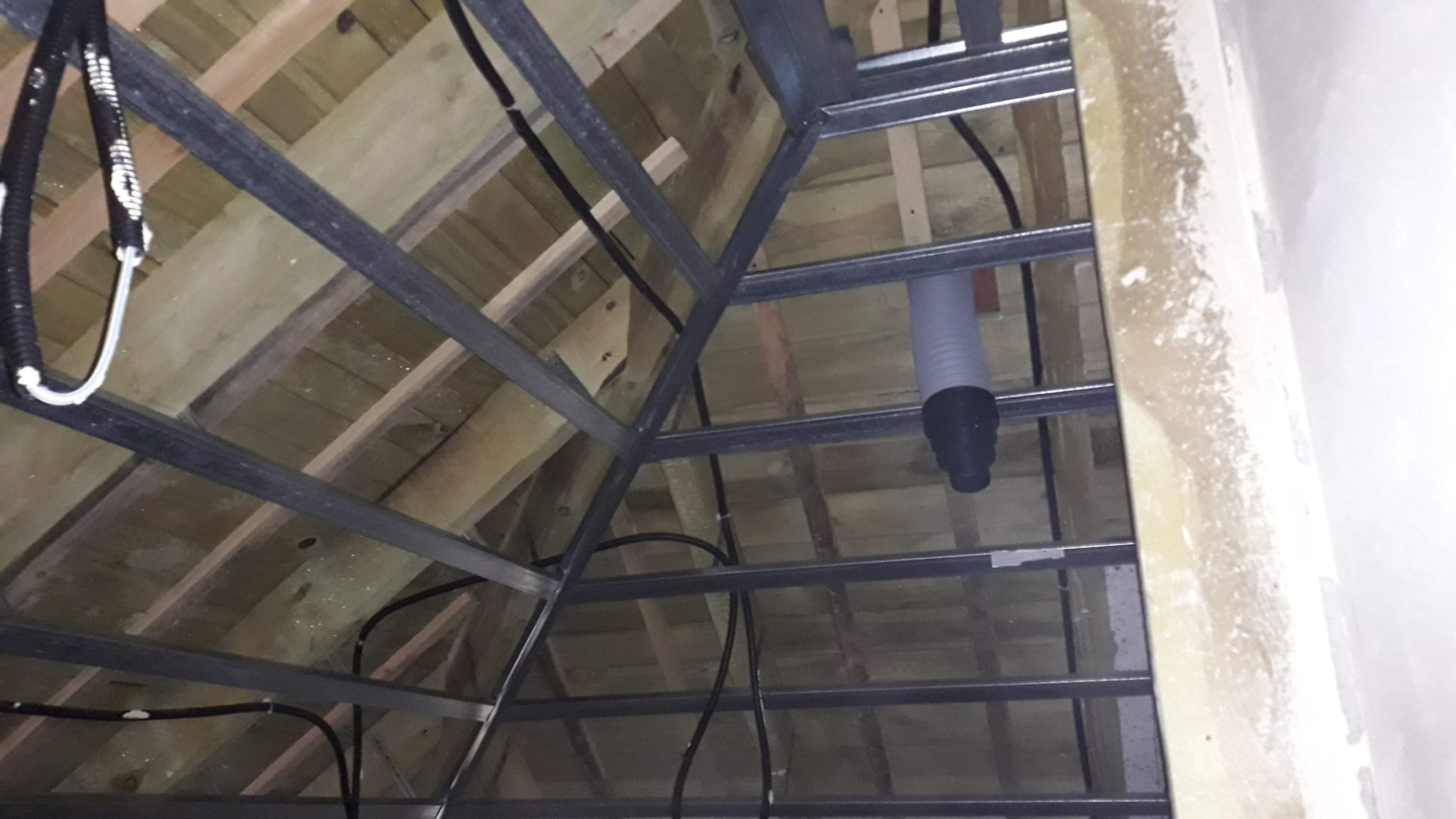 Konstrukcja metalowa podwieszana do drewnianego stropu na poddaszu, widoczne przewody elektryczne i rura wentylacyjna, przygotowanie do montażu płyt gipsowo-kartonowych.