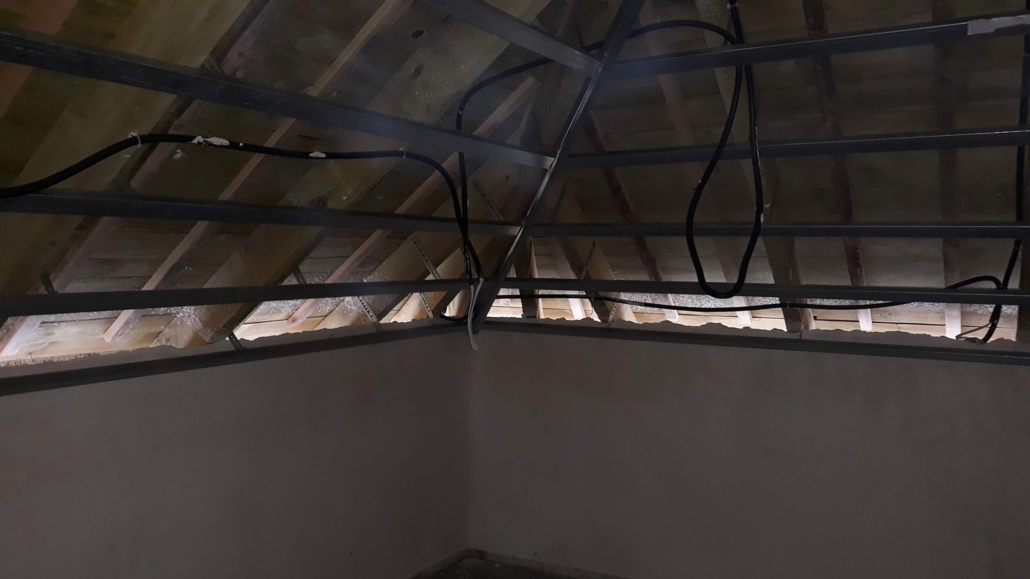 Widok poddasza w trakcie prac: metalowa konstrukcja sufitu podwieszanego, widoczne przewody elektryczne i ocieplenie między krokwiami dachu, ściany przygotowane do dalszych prac wykończeniowych.