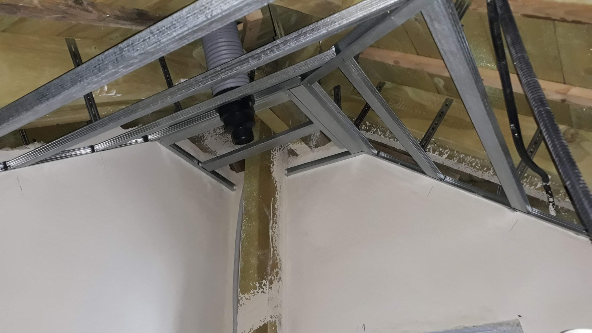 Metalowa konstrukcja sufitu podwieszanego zamontowana pod drewnianym stropem, widoczny przewód wentylacyjny i instalacja elektryczna, ściany częściowo pokryte masą szpachlową.