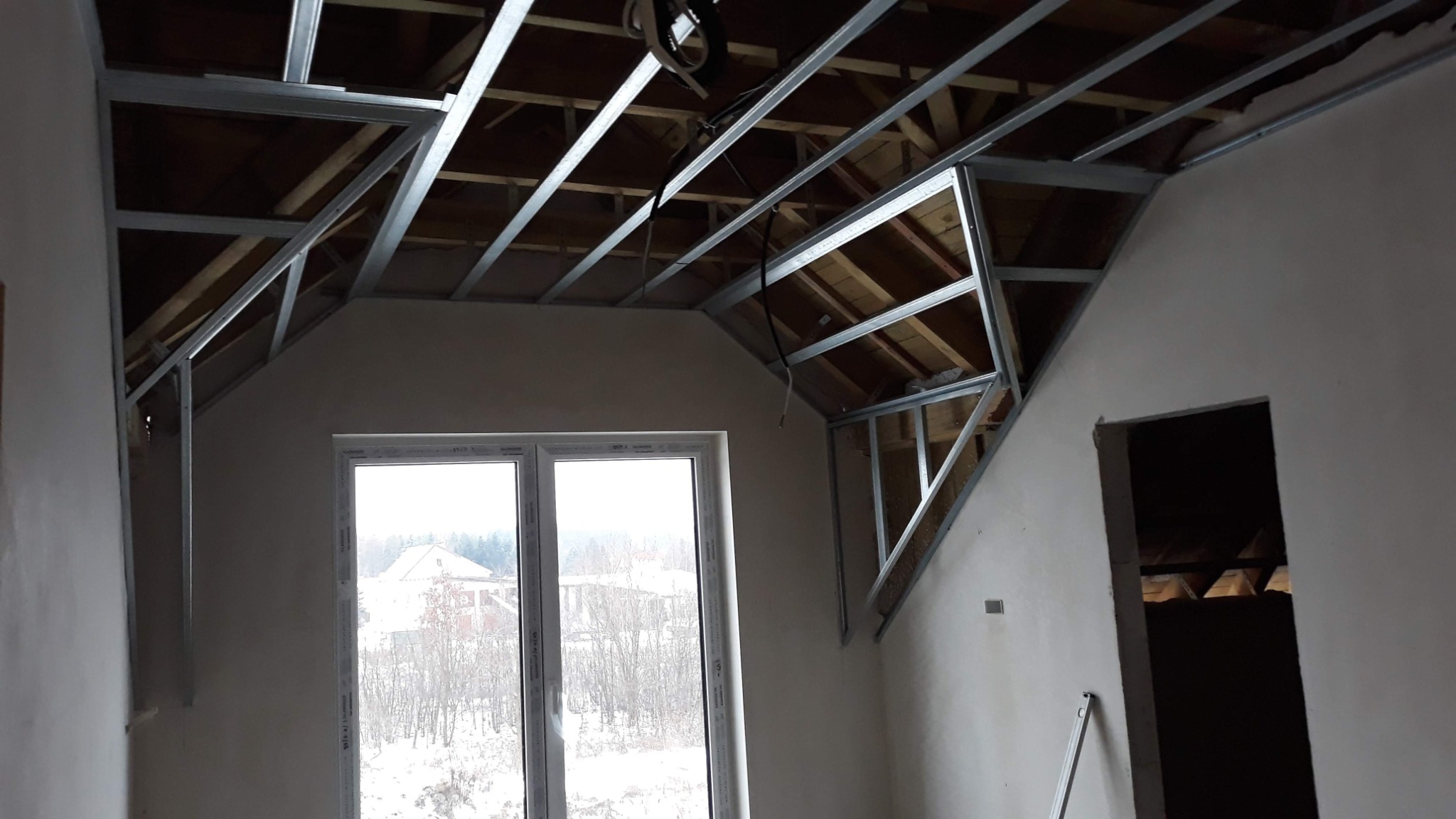 Stelaż z profili metalowych zamontowany pod skośnym dachem na poddaszu, przygotowany do montażu płyt. Widoczne okno dachowe i surowe ściany.