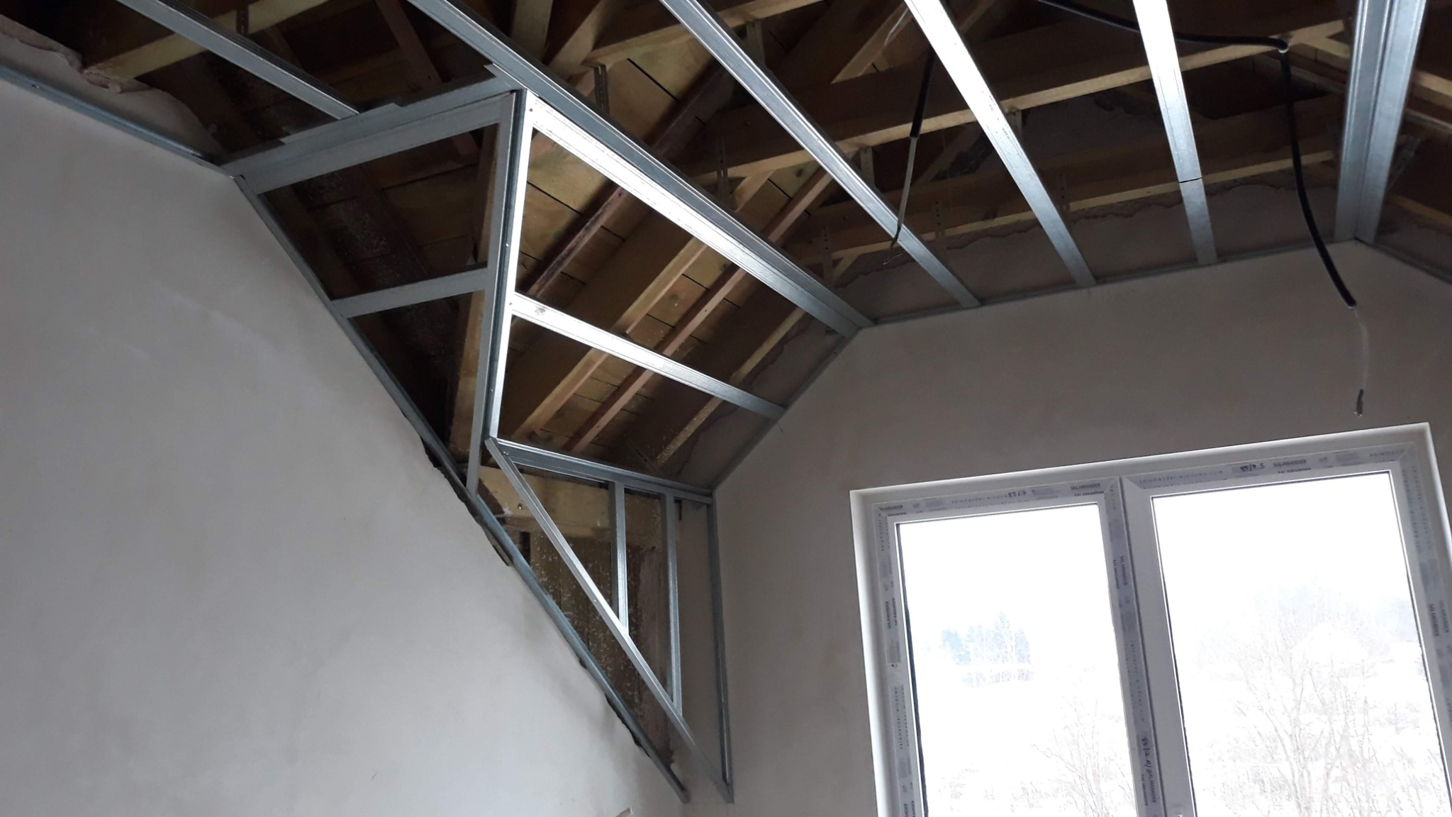 Skośny sufit poddasza w trakcie montażu stelaża z profili metalowych pod płyty gipsowo-kartonowe, widoczne drewniane elementy konstrukcji dachu i przewody elektryczne.