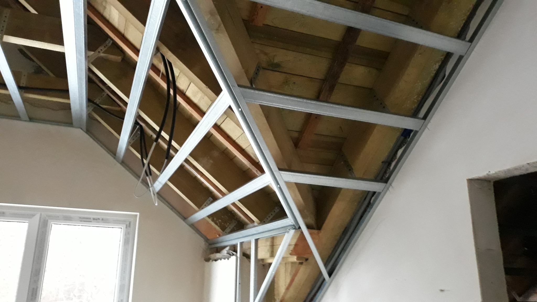 Konstrukcja metalowa podwieszanego sufitu na poddaszu, widoczne drewniane elementy więźby dachowej, kable elektryczne i fragment okna.