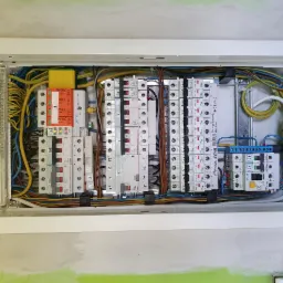 Otwarta skrzynka elektryczna z zamontowanymi bezpiecznikami, ogranicznikami przepięć Simtec i Eaton, oraz uporządkowanym okablowaniem, widoczna na tle surowej ściany przed wykończeniem.