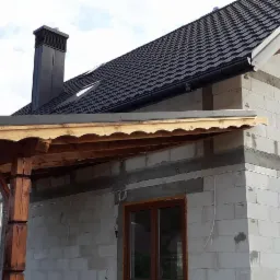 Dom w trakcie budowy z ciemną dachówką, widoczne rynny i obróbki blacharskie, drewniana konstrukcja z dekoracyjnym wykończeniem na tarasie.