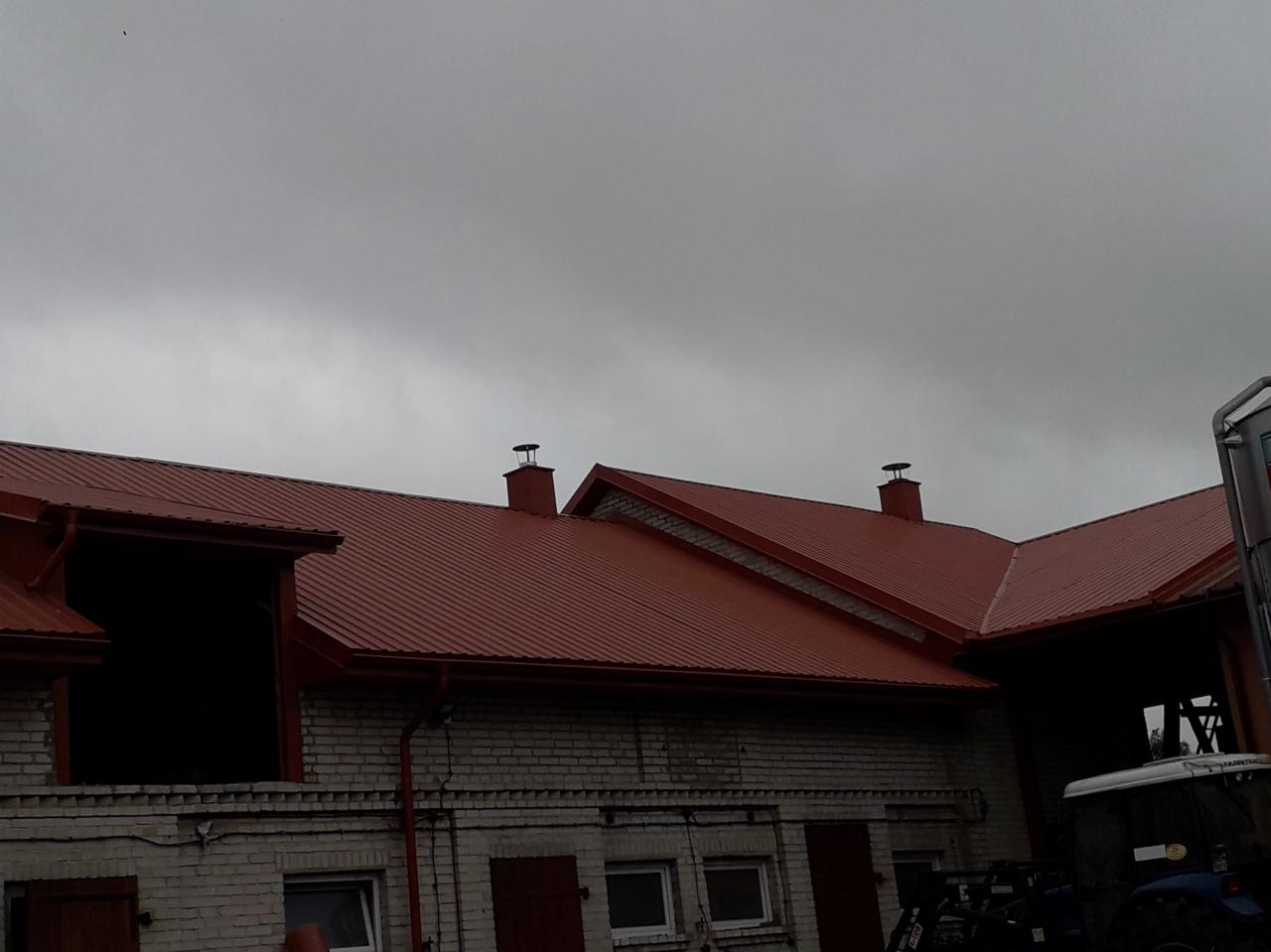 Czerwony dach z blachy trapezowej na budynku z białej cegły, widoczne kominy i rynny, pochmurne niebo.