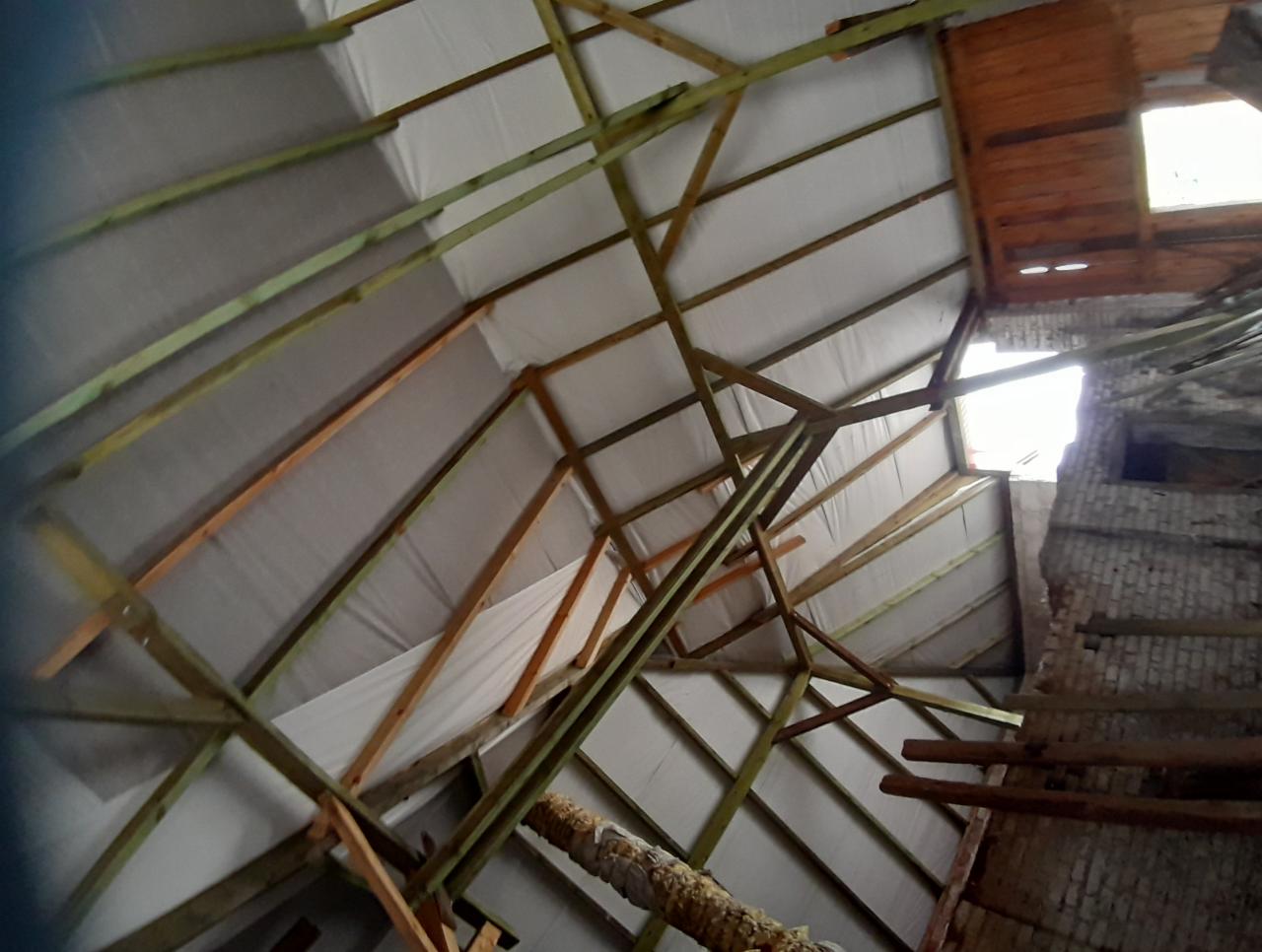 Wnętrze dachu w trakcie budowy z widoczną konstrukcją drewnianą, izolacją i fragmentem murowanego komina z uszkodzeniami.