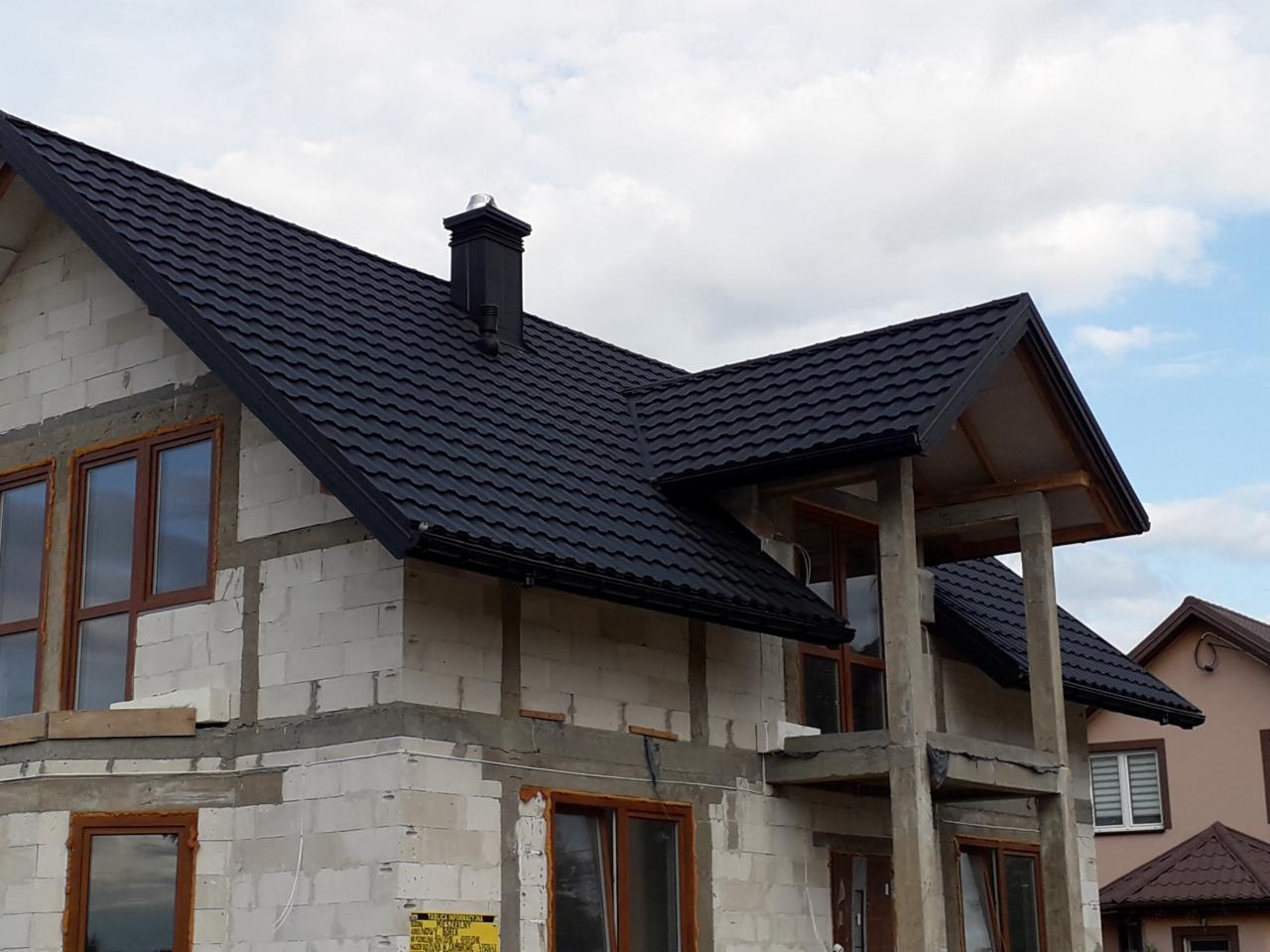 Dom w trakcie budowy z zamontowanym czarnym dachem z blachodachówki i rynnami, widoczne okna z brązowymi ramami i komin z metalowym zakończeniem.
