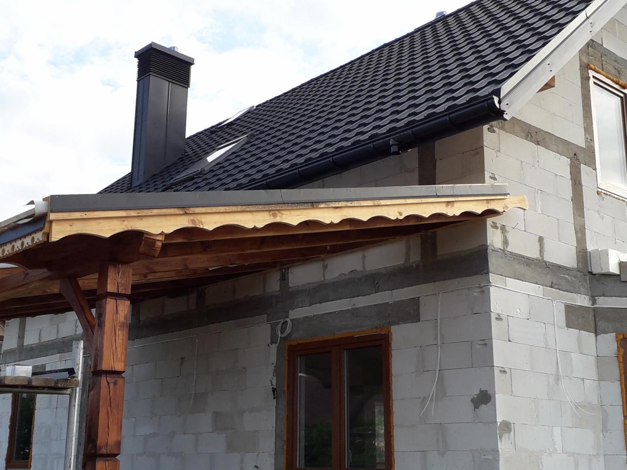 Dom w trakcie budowy z ciemną dachówką, widoczne rynny i obróbki blacharskie, drewniana konstrukcja z dekoracyjnym wykończeniem na tarasie.