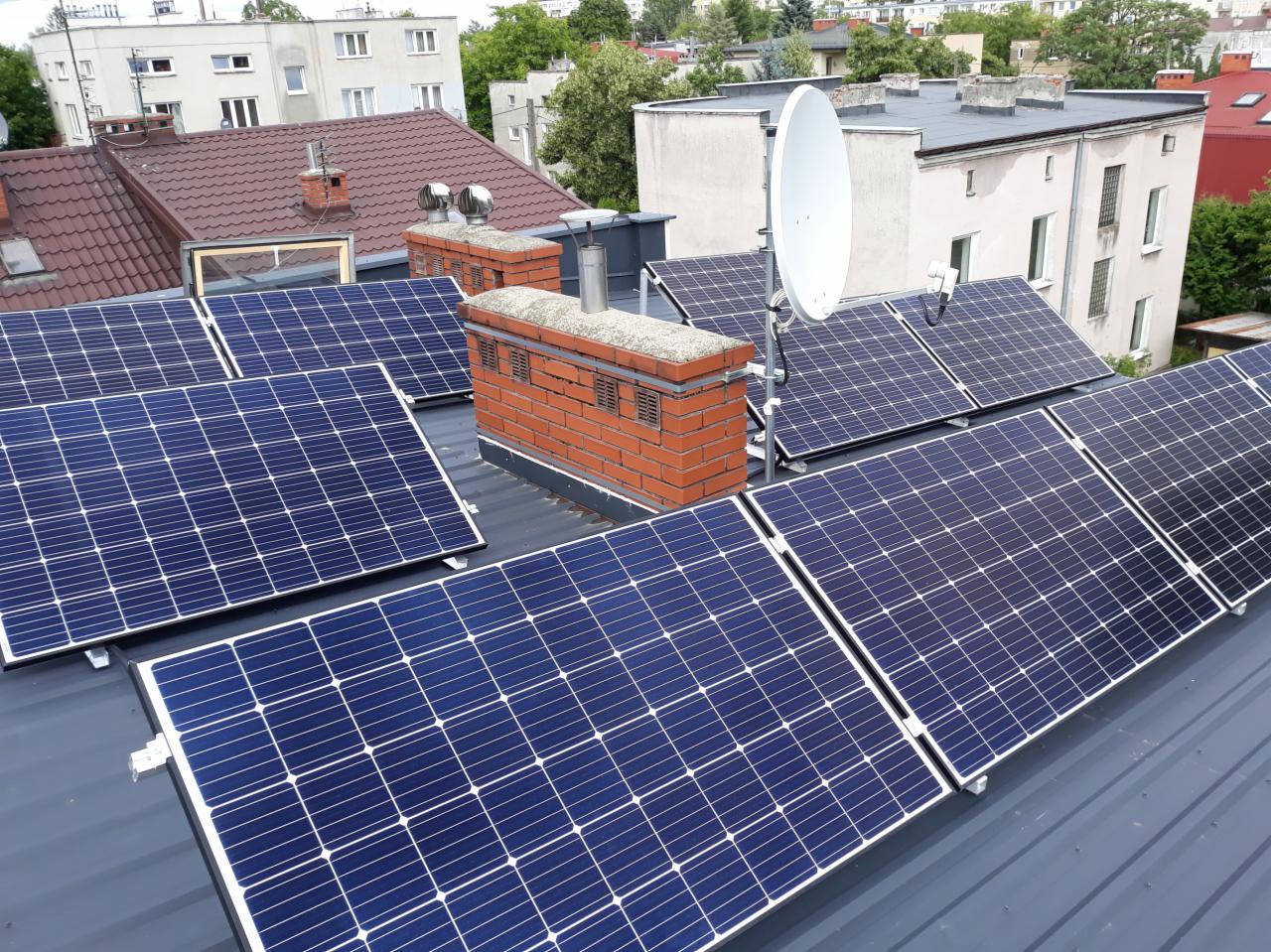 Instalacja paneli słonecznych na dachu budynku w Warszawie, widok z góry, z kominem i anteną satelitarną w tle.