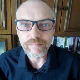 Portret mężczyzny w okularach i czarnej koszuli, z książkami widocznymi w tle.