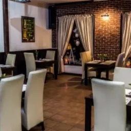 Wnętrze restauracji z ciemnymi drewnianymi stołami, białymi krzesłami, ceglaną ścianą i oknami z zasłonami, dekorowane balonami i lampkami.