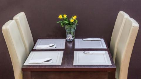 Stół dla czterech osób z kremowymi krzesłami, nakryty szarymi podkładkami i białymi serwetkami, wazon z żółtymi różami jako dekoracja, ciemnobrązowe tło.