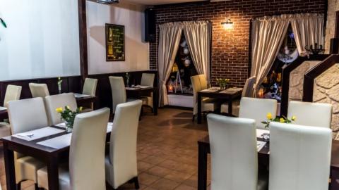 Wnętrze restauracji z ciemnymi drewnianymi stołami, białymi krzesłami, ceglaną ścianą i oknami z zasłonami, dekorowane balonami i lampkami.