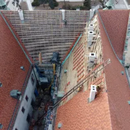 Widok z góry na remontowany dach budynku z czerwonej dachówki, widoczne rusztowania, drewniana konstrukcja nośna oraz pracownicy w trakcie prac dekarskich.