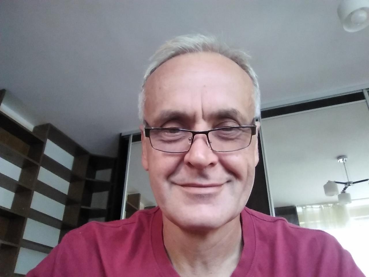 Portret uśmiechniętego mężczyzny w okularach, w bordowej koszulce, na tle wnętrza z regałem i lampą.