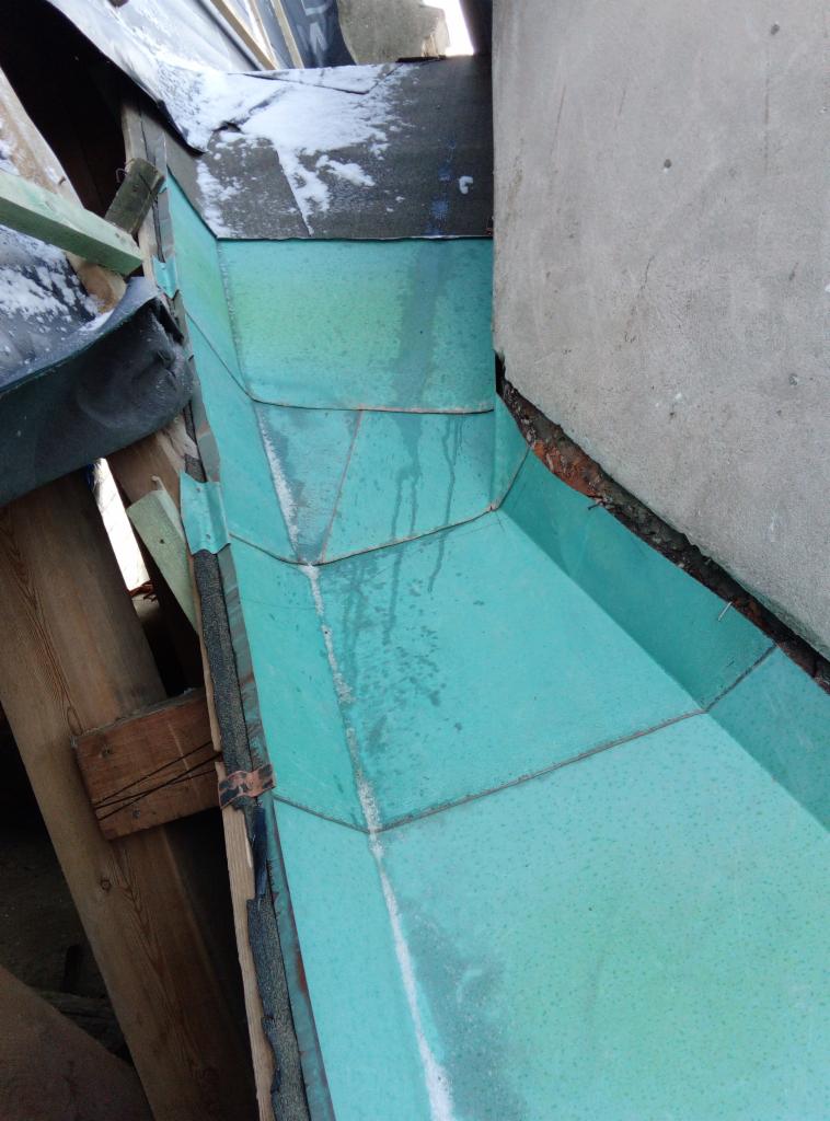 Fragment dachu z miedzianymi panelami w kolorze patynowanej zieleni, przylegający do betonowej ściany, widoczne drewniane elementy konstrukcji.