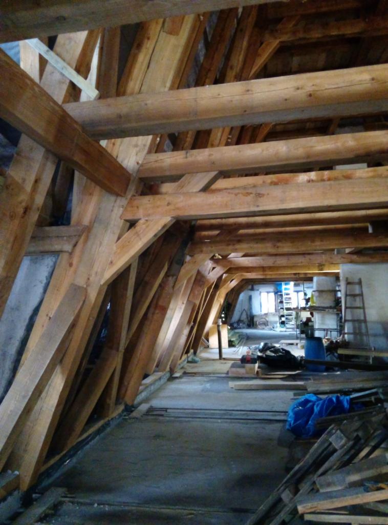 Wnętrze poddasza z drewnianą konstrukcją więźby dachowej w trakcie montażu, widoczne elementy nośne i podpierające, zarys okna w tle, narzędzia i materiały budowlane na podłodze.