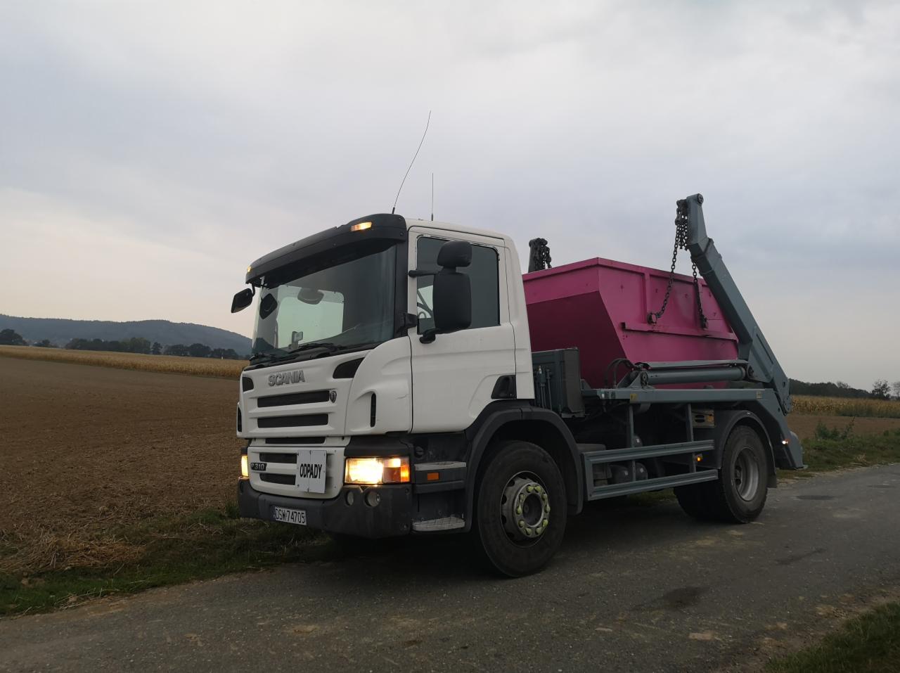 Biała ciężarówka Scania z różowym kontenerem na odpady, zaparkowana na poboczu drogi polnej przy zachmurzonym niebie.