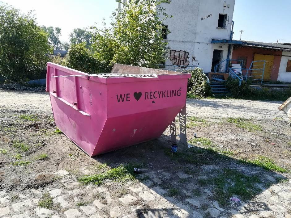 Różowy kontener na gruz z napisem 'WE ❤️ RECYKLING' stoi na placu budowy, w tle budynek z graffiti.