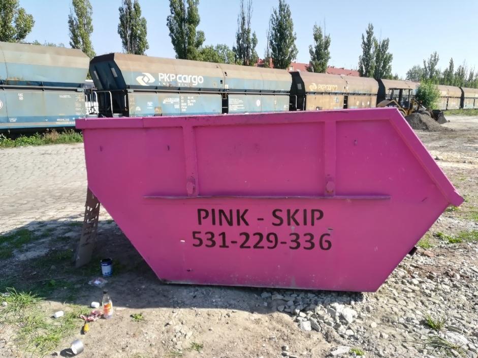 Różowy kontener na gruz z napisem 'PINK - SKIP' i numerem telefonu na tle wagonów PKP Cargo i koparki.