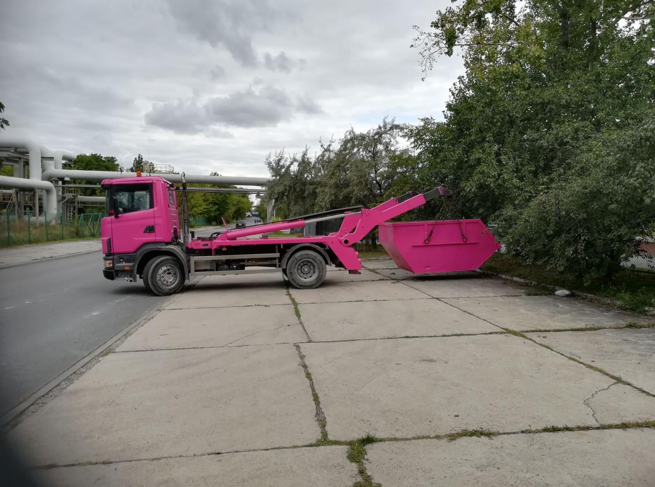 Różowy hakowiec z kontenerem na betonowej nawierzchni, w tle instalacje przemysłowe i zieleń.