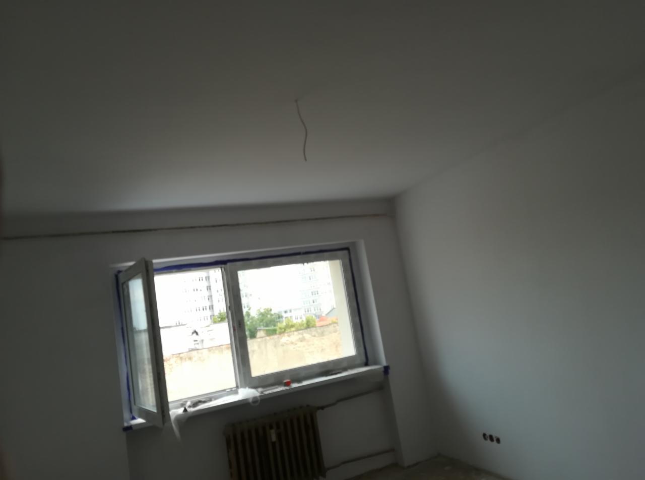 Wnętrze pokoju po świeżym pomalowaniu na biało, z zabezpieczonym taśmą oknem i wystającym kablem z sufitu.