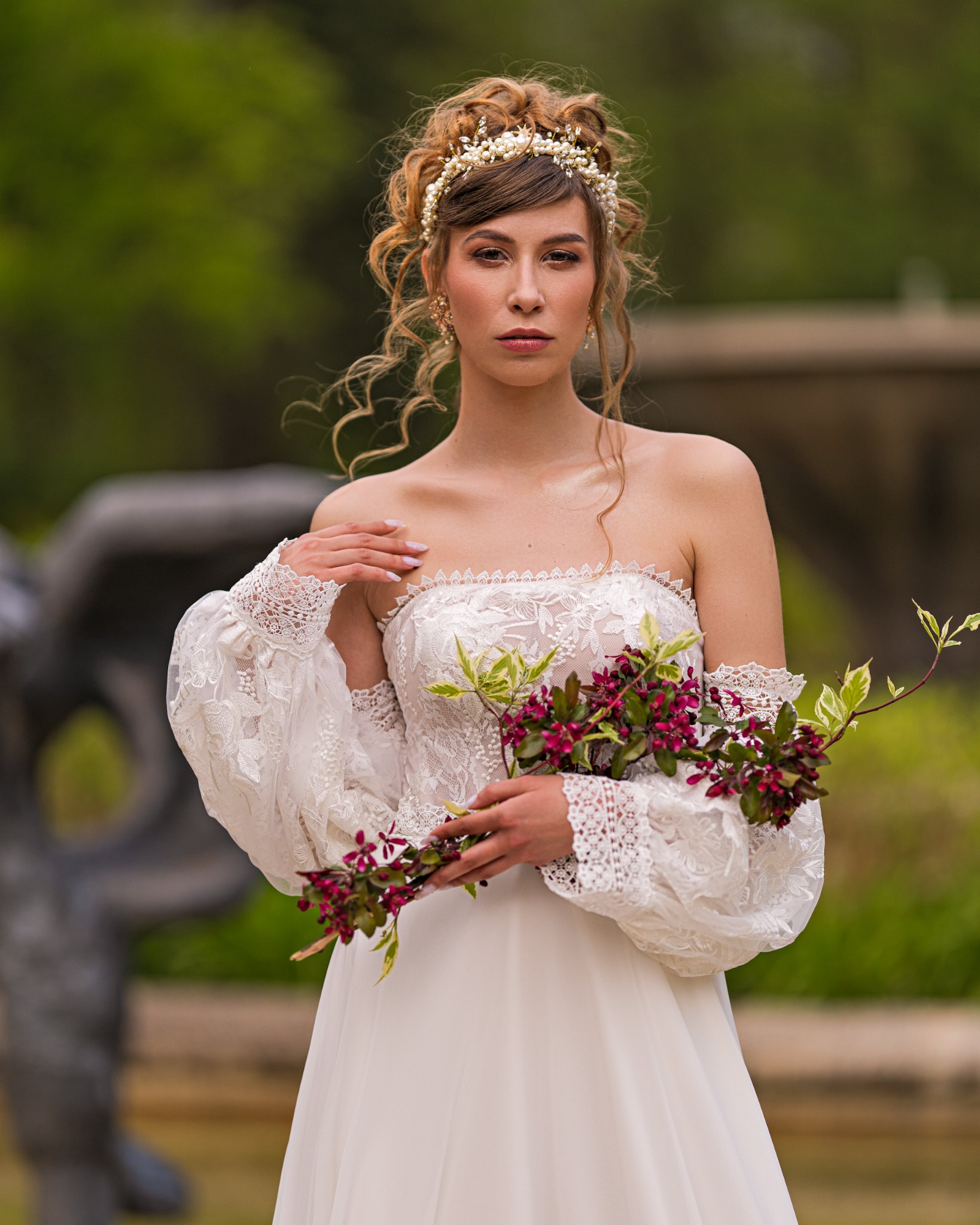 Panna młoda z delikatnym upięciem włosów, perłową ozdobą i bukietem w rękach, w koronkowej sukni bez ramiączek na tle zieleni i rzeźby.